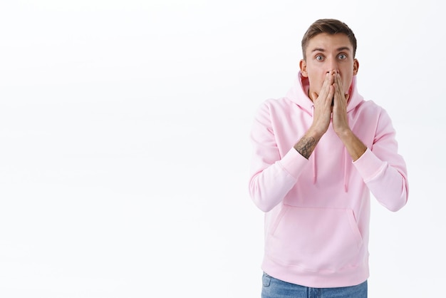 Shocked young blond guy react speechless to astounding news gossips gasping cover mouoth and stare with disbelief at camera popped eyes impressed standing over white background