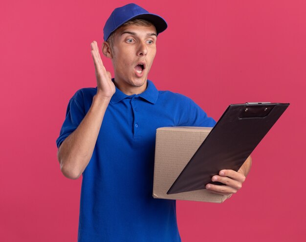 Shocked young blonde delivery boy stands with raised hand and holds cardbox looking at clipboard isolated on pink wall with copy space