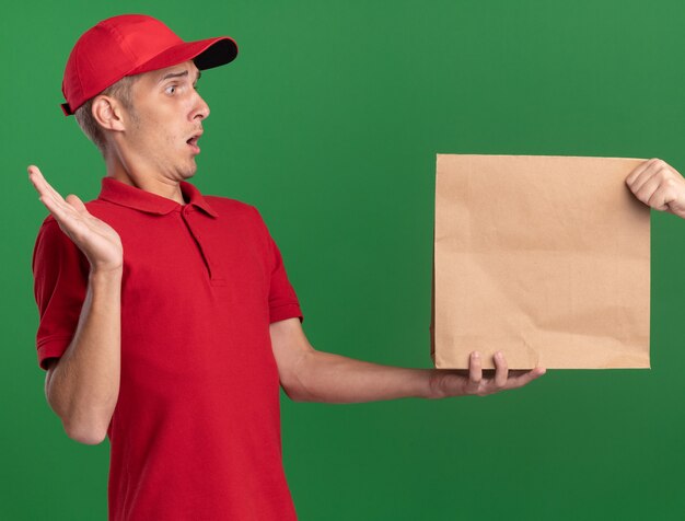Shocked young blonde delivery boy stands with raised hand and takes paper package on green 