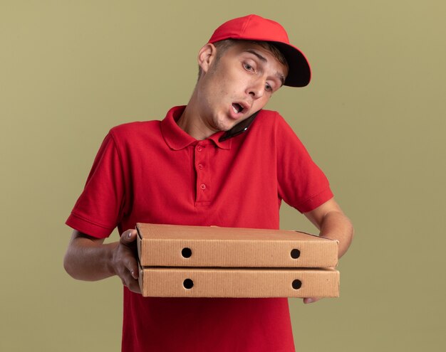Shocked young blonde delivery boy talks on phone holding and looking at pizza boxes isolated on olive green wall with copy space