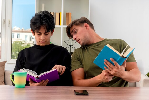 Shocked young blonde handsome man holds book sitting at table and looking at book of pleased young brunette handsome guy inside living room