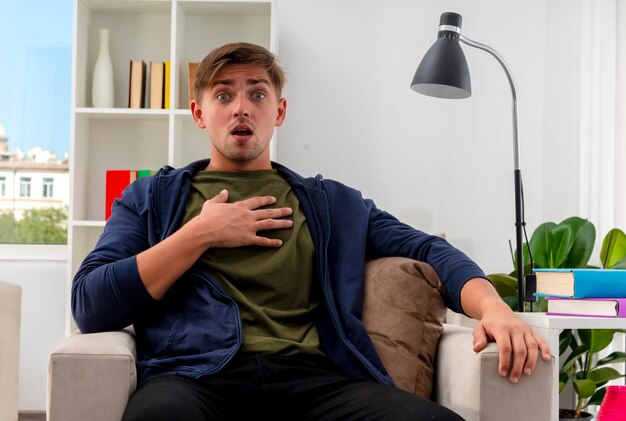 Shocked young blonde handsome man sits on armchair putting hand on chest inside the living room