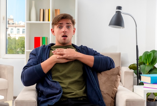 Shocked young blonde handsome man sits on armchair putting hands on chest looking at camera inside living room