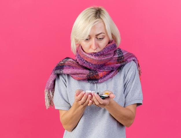 Shocked young blonde ill slavic woman wearing scarf holds and looks at packs of medical pills