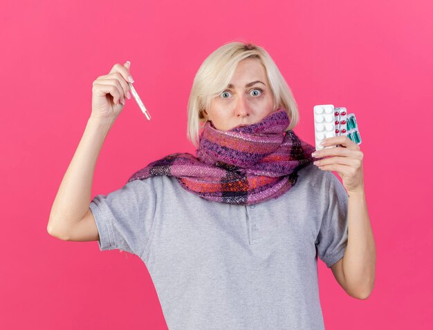 Shocked young blonde ill slavic woman wearing scarf holds packs of medical pills