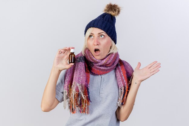 Shocked young blonde ill slavic woman wearing winter hat and scarf stands with raised hand and holds medicine in glass bottle isolated on white wall with copy space