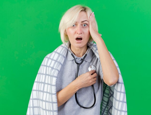 Shocked young blonde ill slavic woman wrapped in plaid puts hand on head and holds stethoscope isolated on green wall with copy space