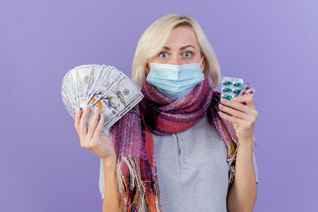 医療マスクとスカーフを身に着けているショックを受けた若い金髪の病気の女性は、紫色の壁に隔離されたお金と医療薬のパックを保持します