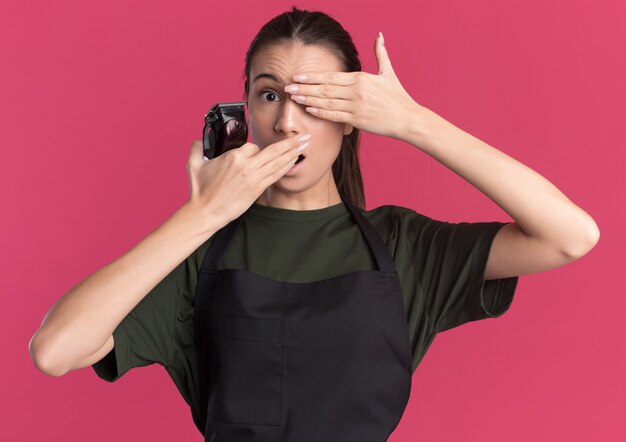 Shocked young brunette barber girl in uniform cobers eye and mouth holding hair clippers