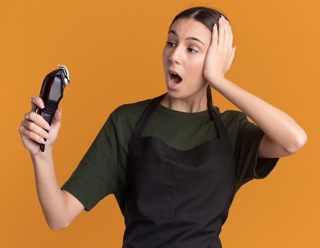 Shocked young brunette barber girl in uniform puts hand on head holding and looking at hair clippers isolated on orange wall with copy space