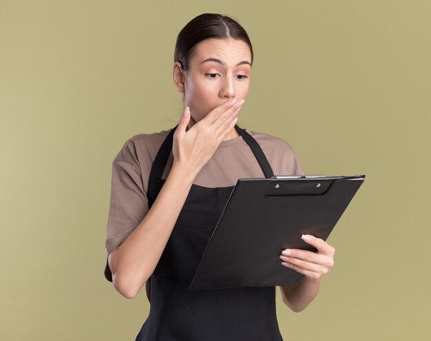 Shocked young brunette barber girl in uniform puts hand on mouth holding and looking at clipboard isolated on olive green wall with copy space