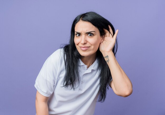 Shocked young brunette caucasian girl holds hand close to ear isolated on purple wall