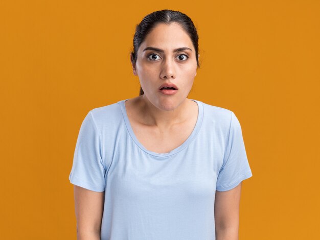 Shocked young brunette caucasian girl looking at camera on orange