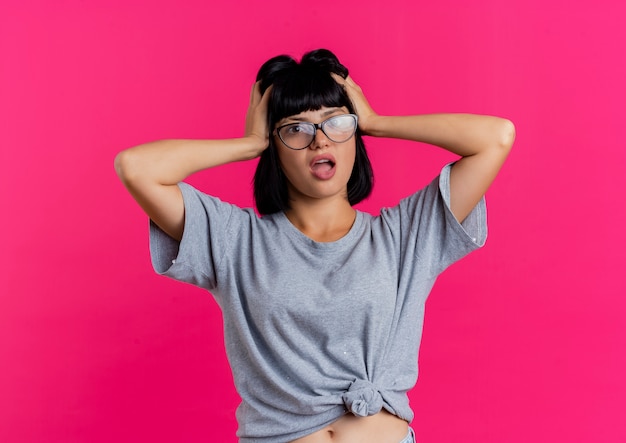 Free photo shocked young brunette caucasian girl in optical glasses holds head and looks at camera isolated on pink background with copy space