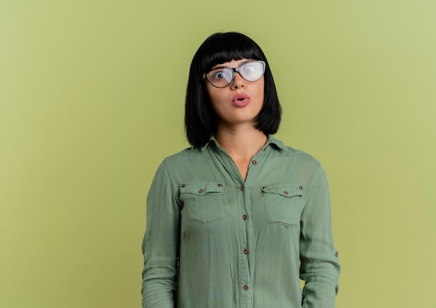 Shocked young brunette caucasian girl in optical glasses looks at camera isolated on olive green background with copy space