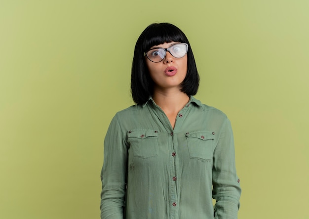 Шокированная молодая брюнетка кавказская девушка в оптических очках смотрит в камеру, изолированную на оливково-зеленом фоне с копией пространства