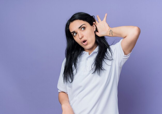 Shocked young brunette caucasian girl puts hand behind ear trying to hear looking at side isolated on purple wall