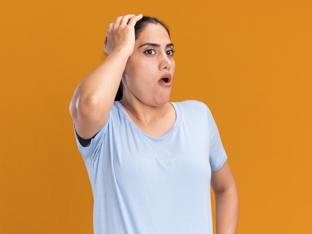 Shocked young brunette caucasian girl puts hand on head and looks at side isolated on orange wall with copy space