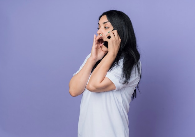 Шокированная молодая брюнетка кавказская девушка стоит боком, разговаривает по телефону, изолированном на фиолетовой стене