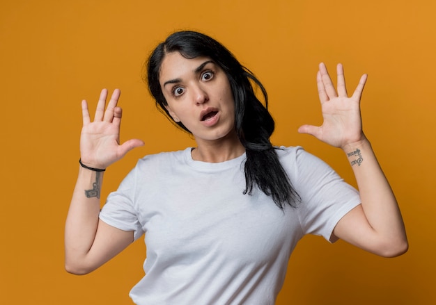 Shocked young brunette caucasian girl stands with raised hands isolated on orange wall