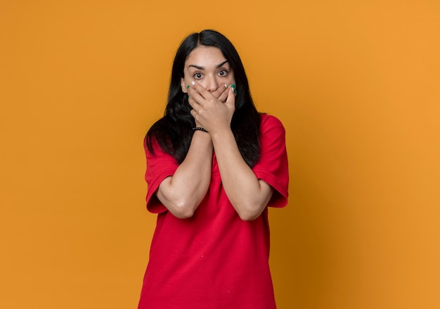 Shocked young brunette caucasian girl wearing red shirt holds mouth with two hands isolated on orange wall