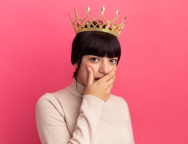 Foto gratuita ragazza caucasica castana giovane scioccata con corona mette la mano sulla bocca e guarda la telecamera sul rosa