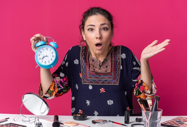 Shocked young brunette girl sitting at table with makeup tools holding alarm clock isolated on pink wall with copy space