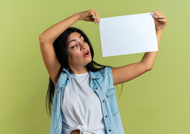 Shocked young caucasian girl holds and looks at paper sheet isolated on olive green background with copy space