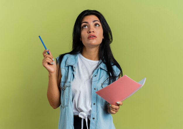 Shocked young caucasian girl holds pen and notebook looking up isolated on olive green background with copy space