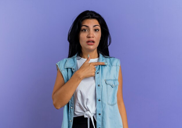 Shocked young caucasian girl points at side looking at camera isolated on purple background with copy space