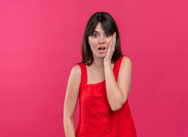 Shocked young caucasian girl puts hand on face and looks at camera on isolated pink background with copy space