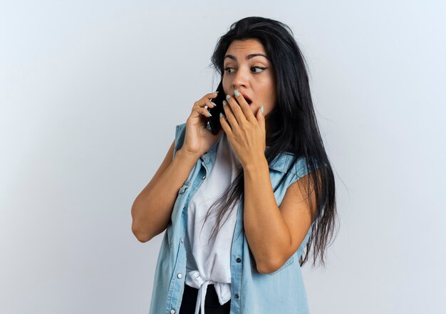 Шокированная молодая кавказская девушка кладет руку на рот, разговаривает по телефону
