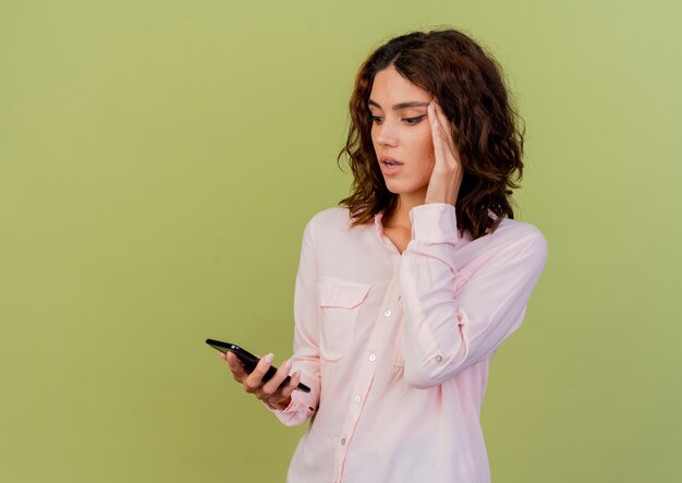 Shocked young caucasian girl puts hand on temple holding and looking at phone isolated on green background with copy space