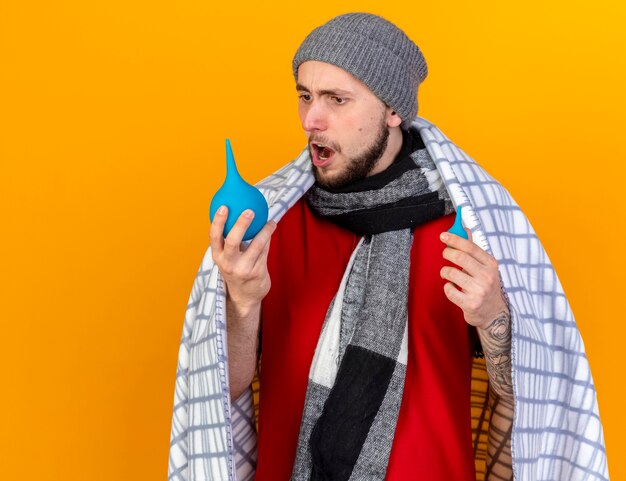 Shocked young caucasian ill man wearing winter hat and scarf wrapped in plaid holds and looks