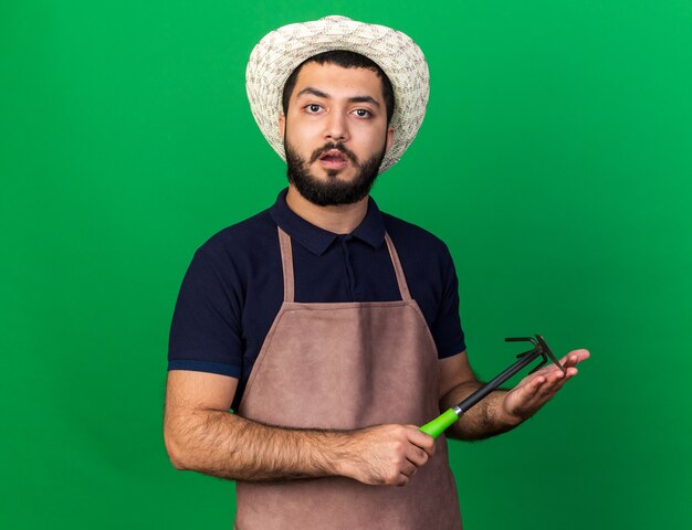 shocked young caucasian male gardener wearing gardening hat holding hoe rake isolated on green wall with copy space