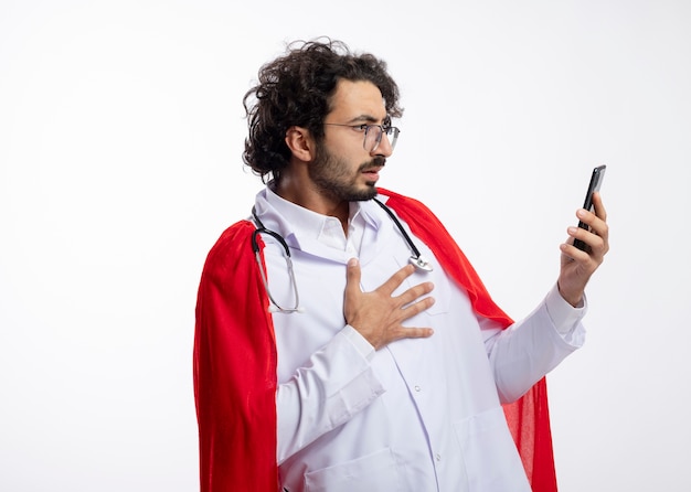 Shocked young caucasian superhero man in optical glasses wearing doctor uniform with red cloak and with stethoscope around neck puts hand on chest and looks at phone with copy space
