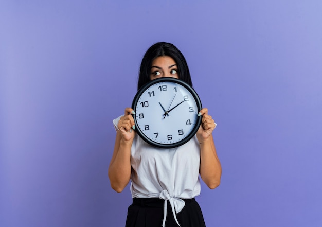 Free photo shocked young caucasian woman holds clock in front of face and looks at side