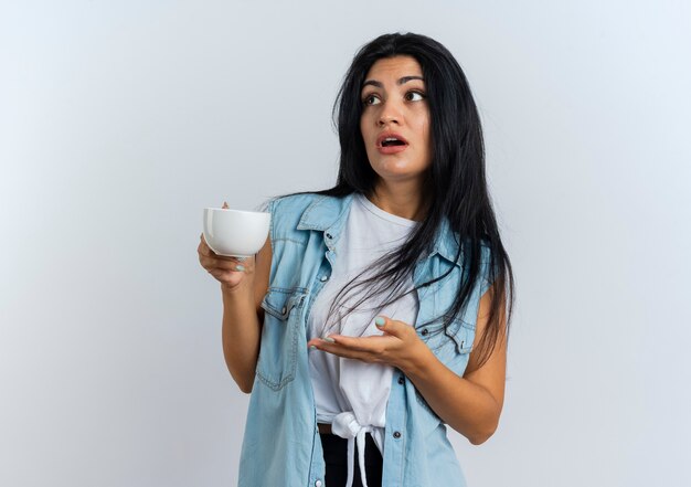 Shocked young caucasian woman holds and points at cup with hand looking at side