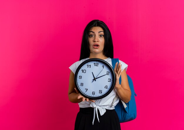 Shocked young caucasian woman wearing backpack holds clock