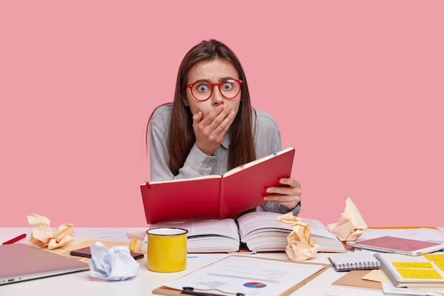 Shocked young college student holds red diary, surrounded with thick opened book, laptop computer, surprised to have deadline task, drinks coffee