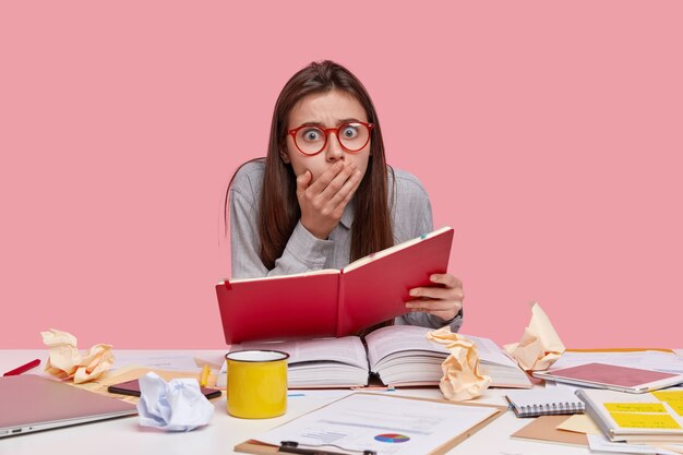 Шокированный молодой студент колледжа держит красный дневник, окруженный толстой открытой книгой, портативный компьютер, удивлен тем, что у него дедлайн задание, пьет кофе
