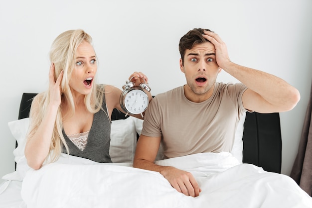 Shocked young couple sitting in bed with opened mouth and holding alarm clock