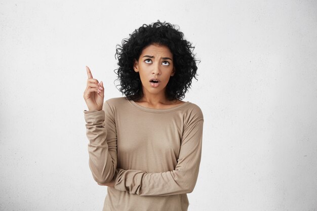 Shocked young dark-skinned woman dressed casually showing something astonishing above her head, keeping mouth wide opened, standing against blank wall