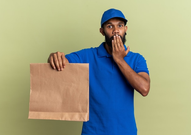 shocked young delivery man putting hand on mouth and holding food package isolated on olive green wall with copy space