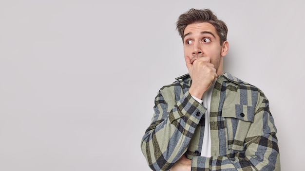 Shocked young European adult man stares bugged eyes covers mouth with hand wears checkered shirt concentrated away blank copy space for your advertisement. Human reactions and emotions concept