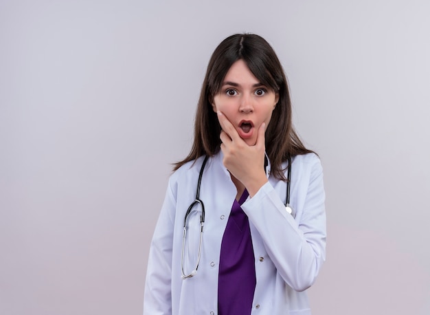 Shocked young female doctor in medical robe with stethoscope puts hand on chin on isolated white background with copy space