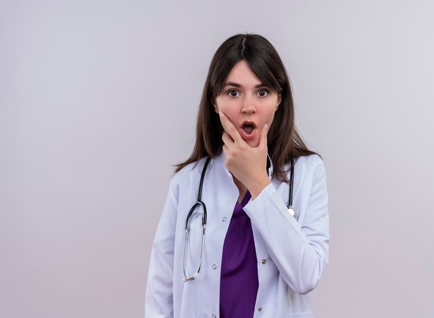 Шокированная молодая женщина-врач в медицинском халате со стетоскопом кладет руку на подбородок на изолированном белом фоне с копией пространства