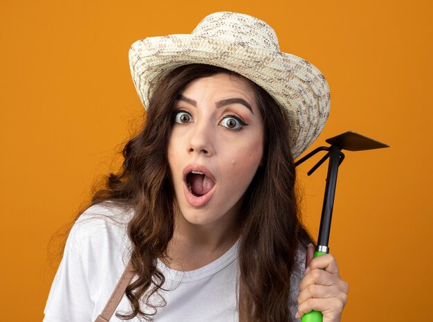 Shocked young female gardener in uniform wearing gardening hat holds hoe rake isolated on orange wall with copy space