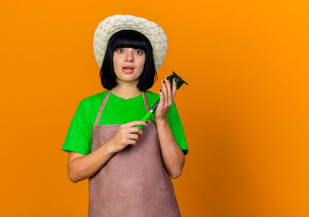 Шокированная молодая женщина-садовник в униформе в садовой шляпе держит грабли для мотыги