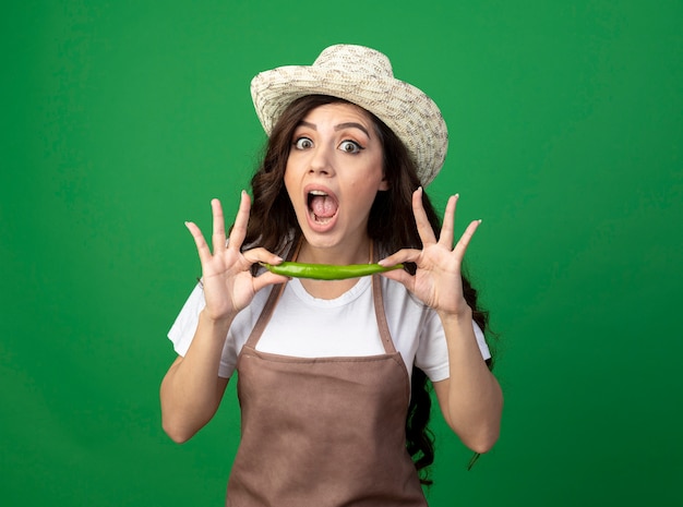 Il giovane giardiniere femminile scioccato in cappello da giardinaggio d'uso uniforme tiene il peperoncino isolato sulla parete verde